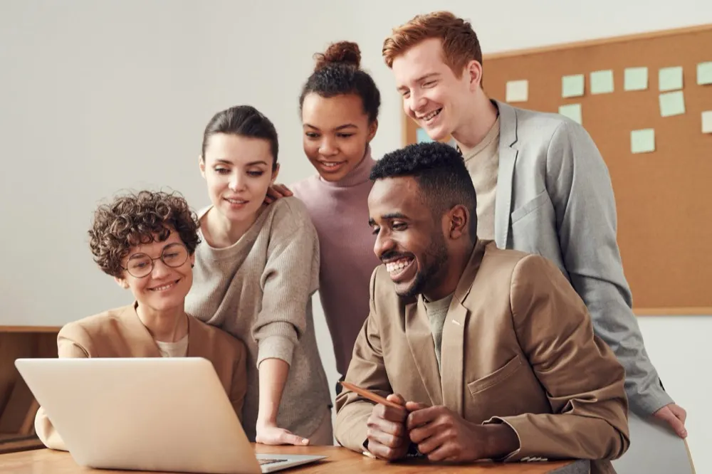 Five coworkers smiling and gathered around a laptop in a bright office