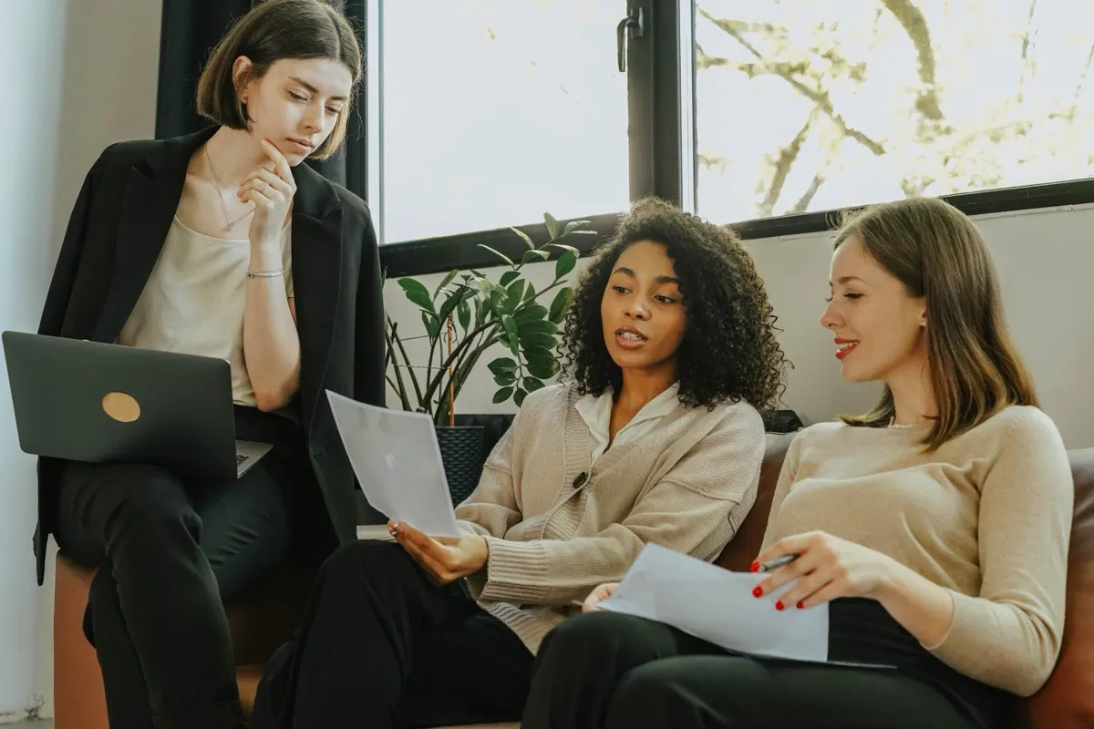Three women sitting together reviewing papers and a laptop