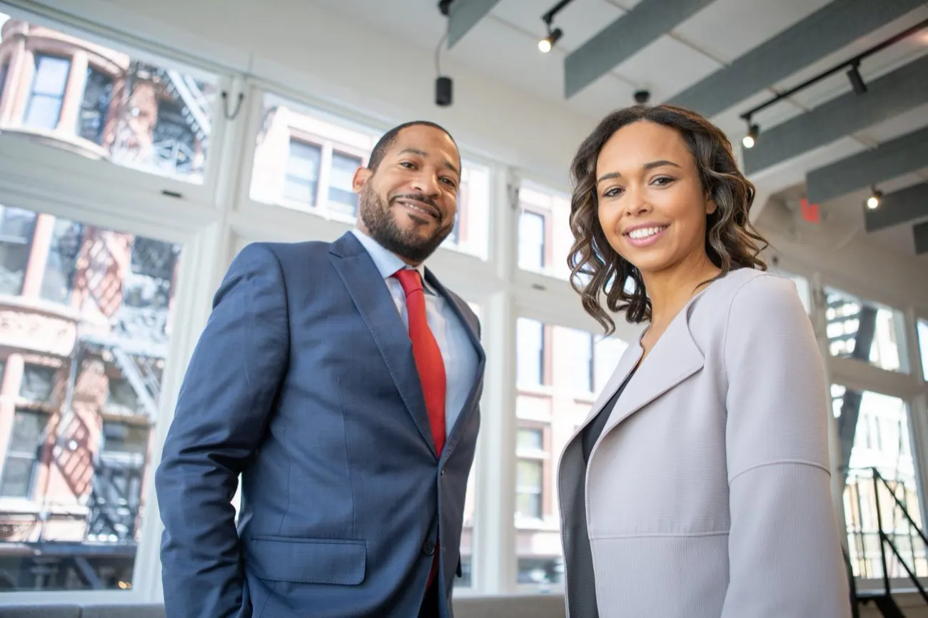 Man in a navy suit and woman in a gray blazer standing side by side in a bright office