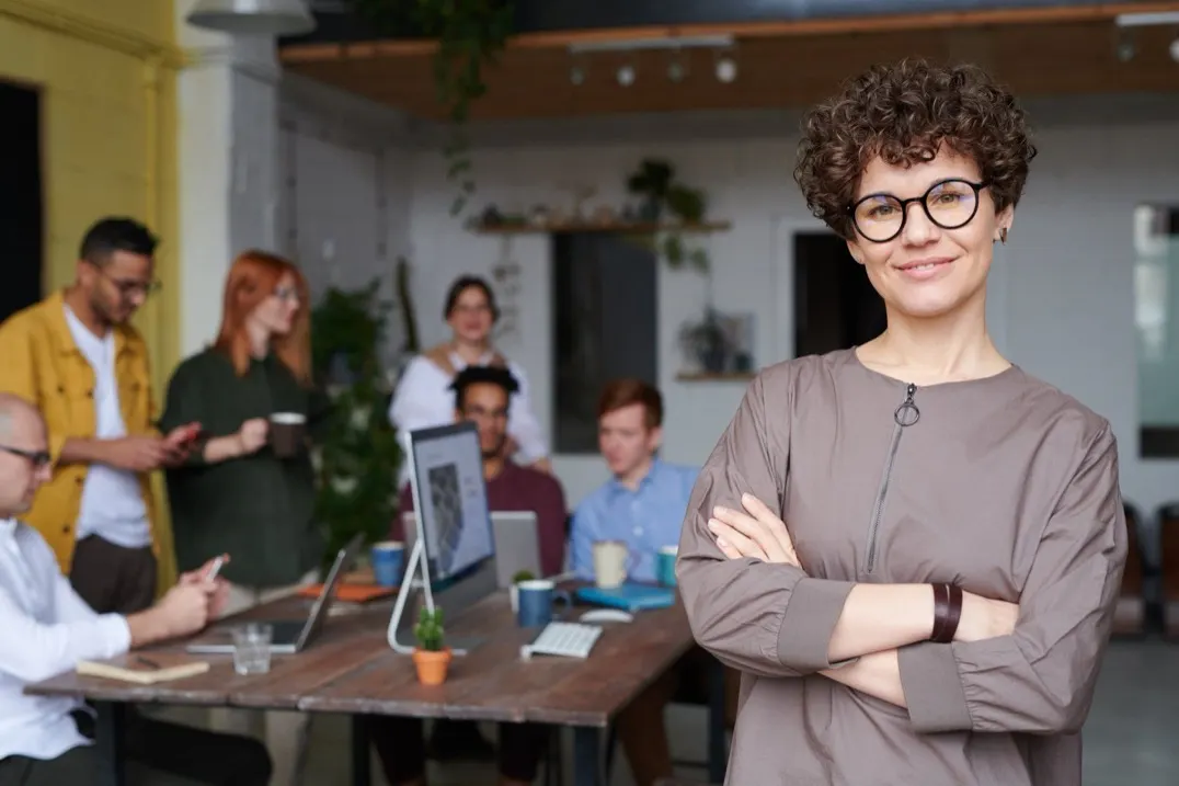 Woman with curly hair and round glasses standing with her arms crossed in front of coworkers at a long table
