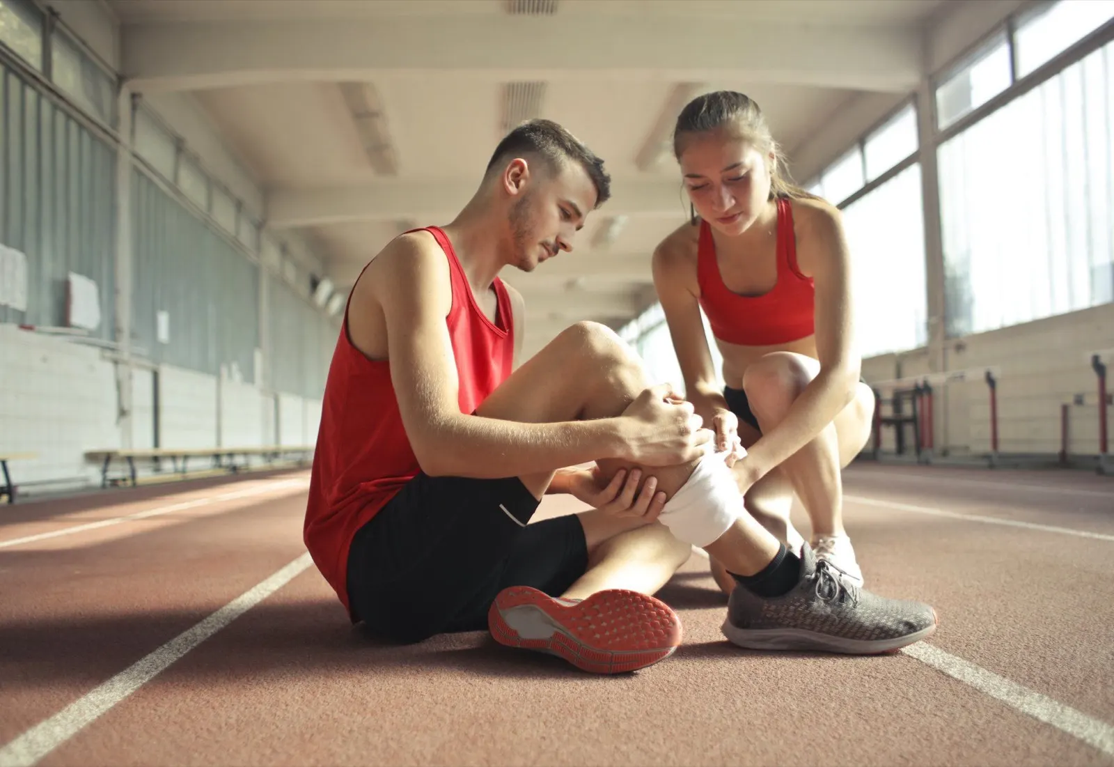 Runner in a red top sitting on an indoor track holding his injured ankle while a woman looks on