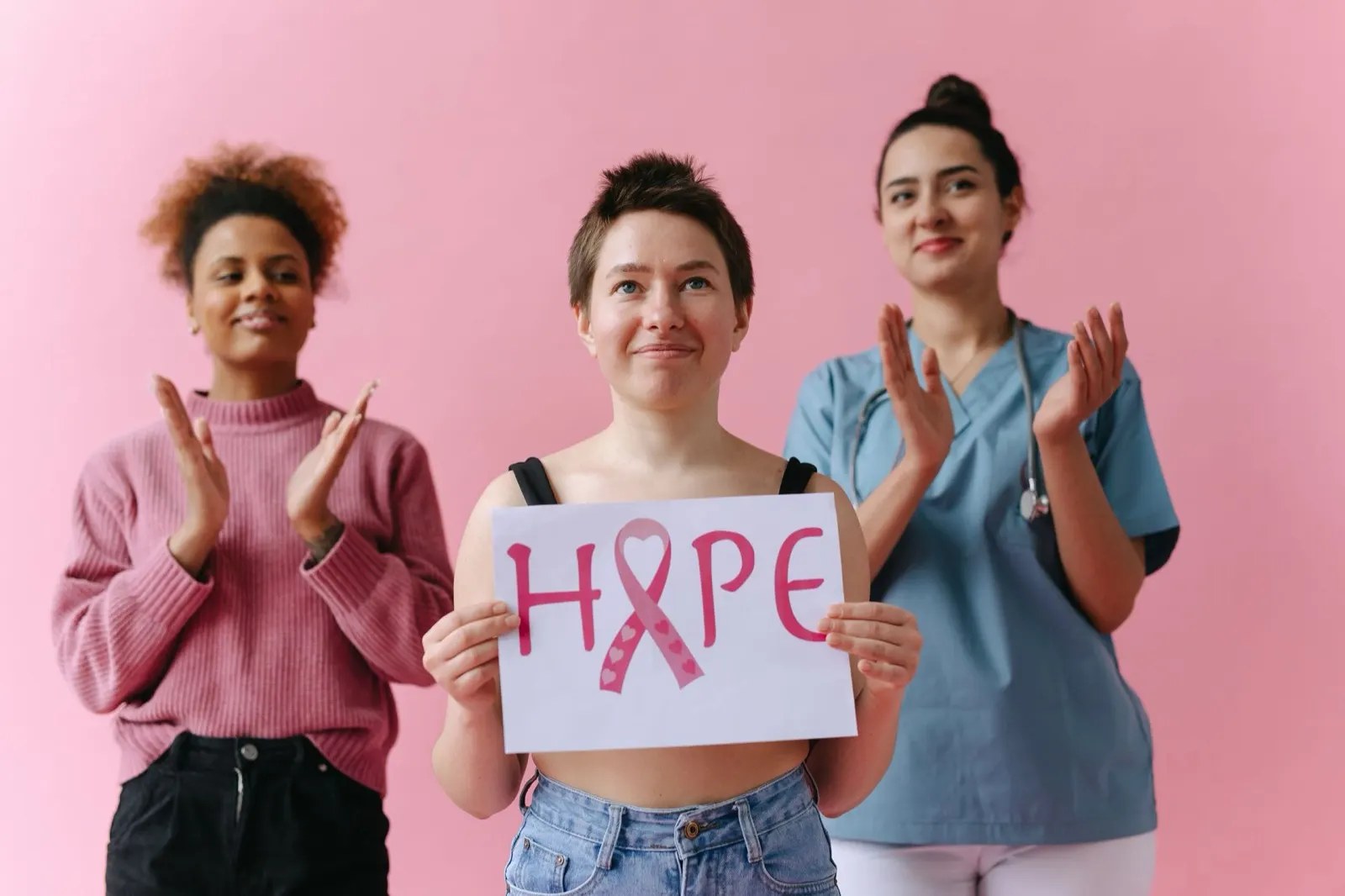 Three women standing against a pink wall, one holding a sign that reads HOPE with a pink ribbon
