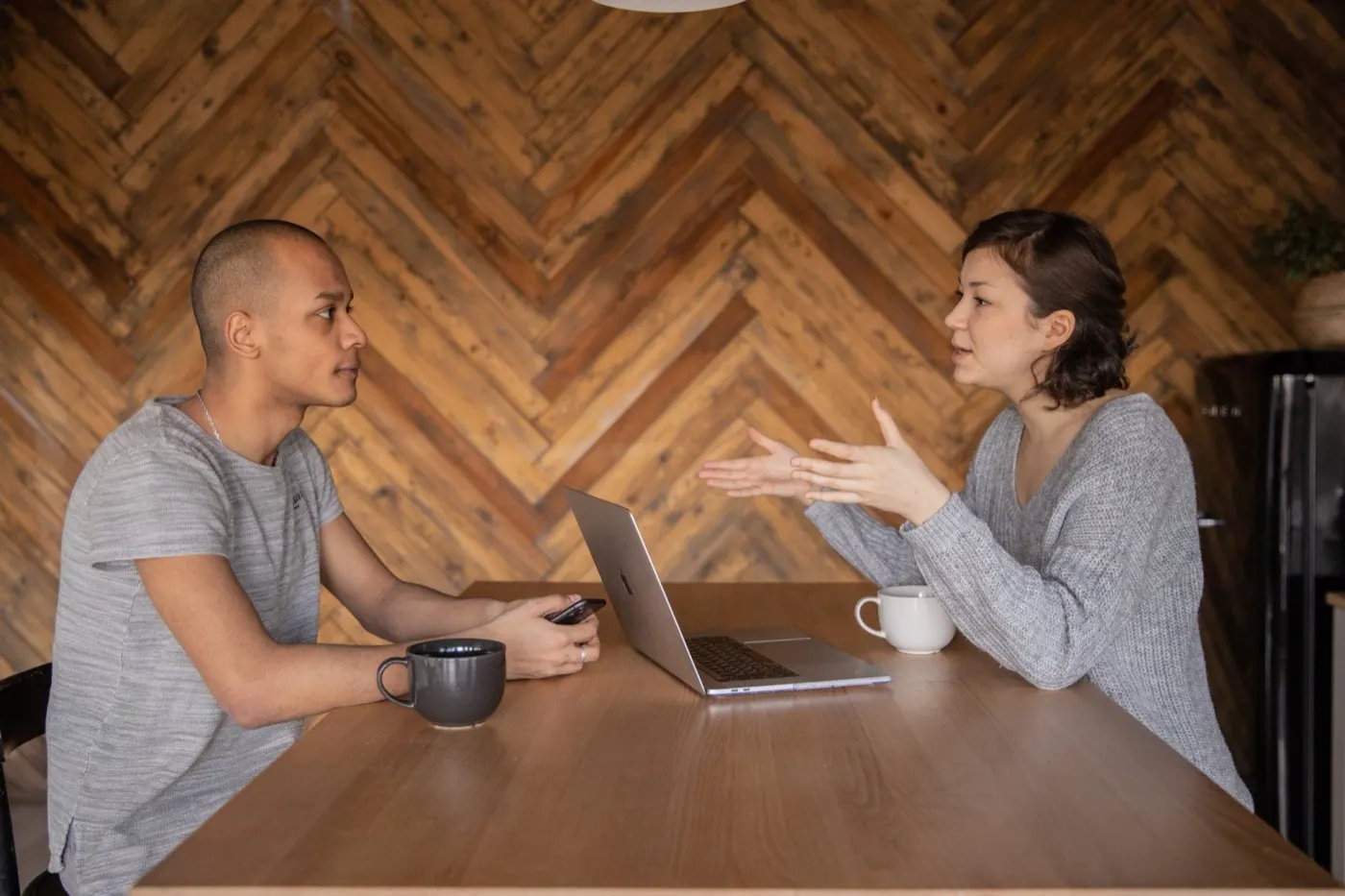 Man and woman talking across a wooden table with a laptop and coffee