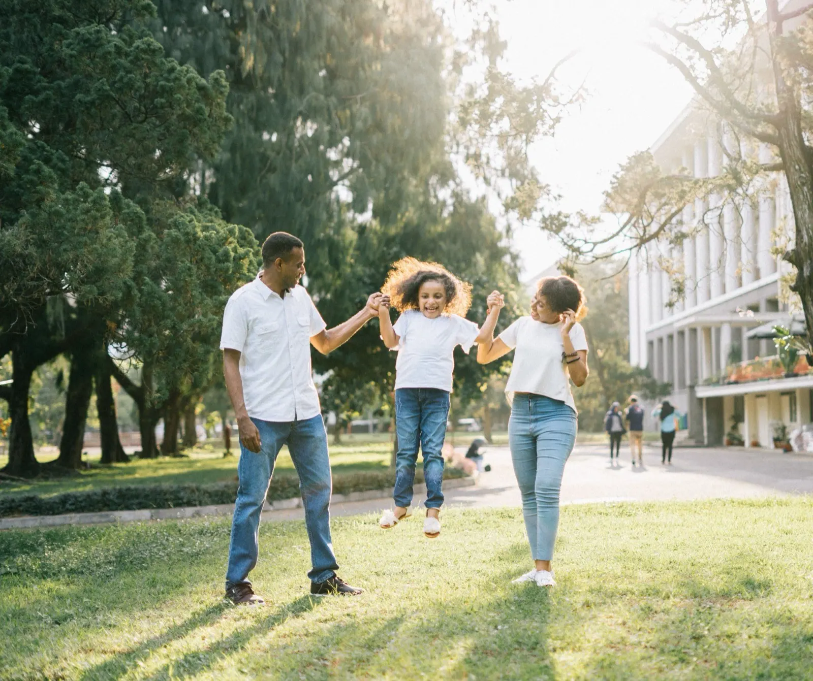 Father and mother swinging their young daughter between them as they walk across a sunny park lawn