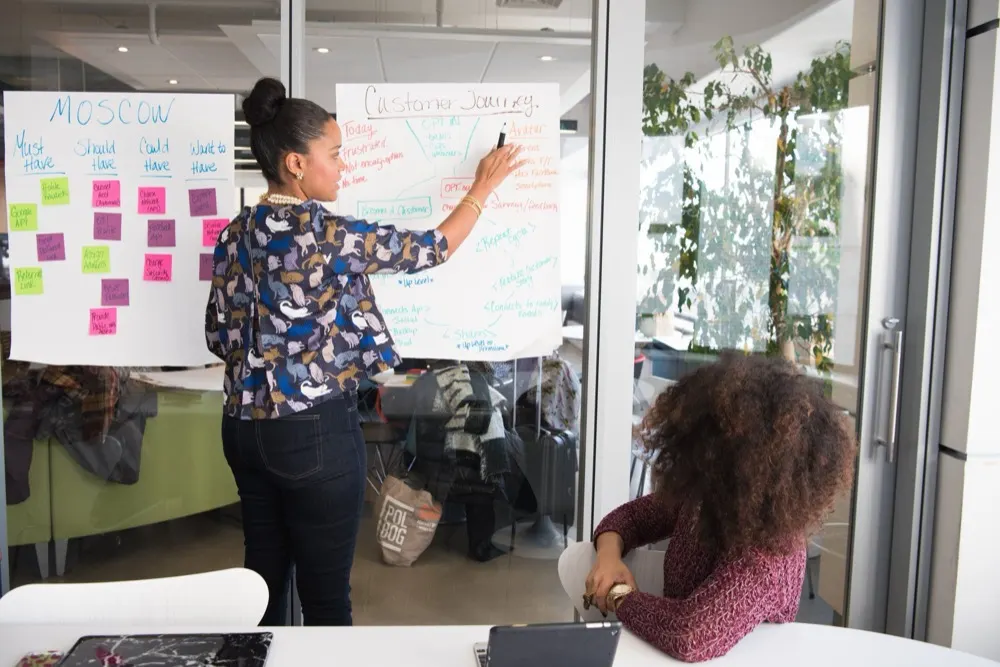 Woman writing ideas on sticky notes on a glass wall while a colleague listens